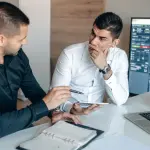 Two businessmen discussing stock market trends with trading screens in an office setting.
