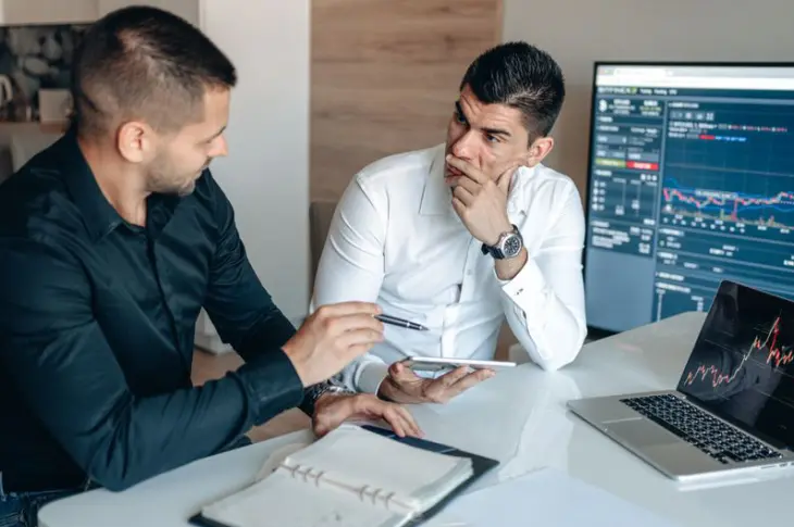 Two businessmen discussing stock market trends with trading screens in an office setting.